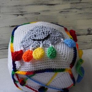 May include: A crocheted cube toy with a rainbow border and a gray cloud with a smiling face in the center. The cloud has colorful pom-poms hanging from it.