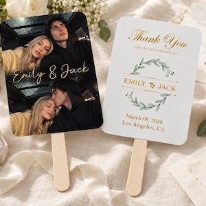 Puede incluir: Dos abanicos de boda rectangulares con mangos de madera. Uno muestra dos fotos de una pareja con el texto dorado Emily & Jack. El otro es blanco con el texto dorado Thank You, Emily & Jack, March 09, 2028, Los Angeles, CA y una corona de hojas verdes.