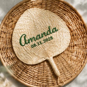May include: A heart-shaped hand fan woven from natural light tan palm leaves, featuring the name Amanda and the date 08.11.2028 embroidered in dark green thread across the center. The fan has a sturdy, wrapped handle with a small loop at the end.