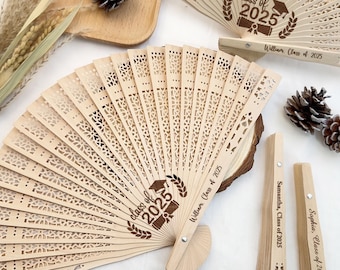 May include: A collection of light brown wooden fans, some open and some closed, with intricate cut-out designs. The fans are engraved with "Class of 2025" and names like "William, Class of 2025". Decorative pinecones are also present.