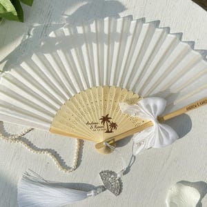 May include: A white folding fan with a light wood handle. The fan is decorated with a white bow and a pearl necklace. The handle is engraved with the names "Andrew & Karen" and the date "09.14.2026".
