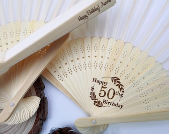 Op de afbeelding: Verschillende open, lichtgekleurde houten waaiers met decoratieve uitsparingen. Eén waaier toont de tekst "Happy 50th Birthday" in een krans. Een andere waaier heeft de tekst "Happy Birthday! Aurora" op het handvat. De waaiers zijn gedeeltelijk open en tonen hun geplooide ontwerp.