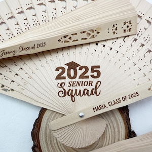 May include: Wooden folding fans with laser-cut designs and the text "2025 Senior Squad." One fan is open, displaying the text, while others are partially visible. The fans are personalized with names and the class year.