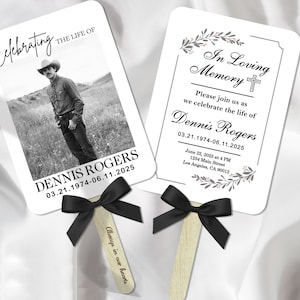 May include: Two white memorial fans with black bows. One fan features a black and white photo of a man in a cowboy hat, with the text "Celebrating the Life of Dennis Rogers." The other fan reads "In Loving Memory."