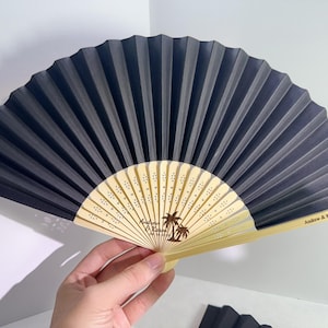 May include: A black and beige folding fan with a palm tree design and the names "Andrew & Karen" along with the date "09.14.2026". The fan is held open, displaying its pleated design.