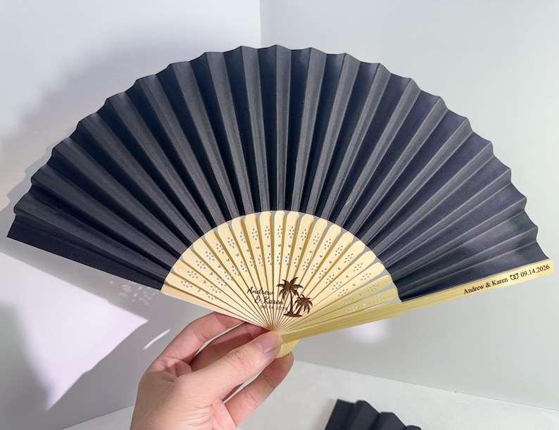 May include: A black and beige folding fan with a palm tree design and the names "Andrew & Karen" along with the date "09.14.2026". The fan is held open, displaying its pleated design.