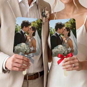 May include: Two rectangular wedding fans featuring a photograph of a couple kissing, with the text CARL & EMILY 07.18.2028 printed in white. One fan has a grey satin ribbon bow, and the other has a red satin ribbon bow attached to the wooden handle.