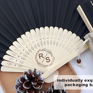 May include: A black and white folding fan with a wooden handle, personalized with initials and a date. The fan is in a beige drawstring bag, with the text "individually exquisite packaging bag".