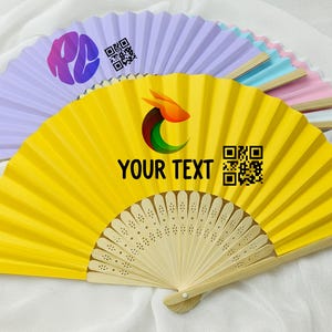 Pode incluir: Uma coleção de leques de papel coloridos, incluindo amarelo, lavanda, rosa e azul. O leque amarelo apresenta o texto "YOUR TEXT" e um código QR. Os leques têm cabos de madeira e estão abertos, mostrando o seu design plissado.