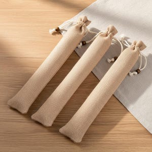 May include: Three beige linen pouches with drawstring closures and wooden bead accents. The pouches are cylindrical and appear to be filled. They are arranged diagonally on a light-colored surface, suggesting a natural or rustic theme.