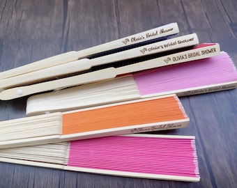 May include: A collection of personalized folding fans in various colors, including pink and orange. The fans have wooden handles and are printed with the text "Olivia's Bridal Shower".