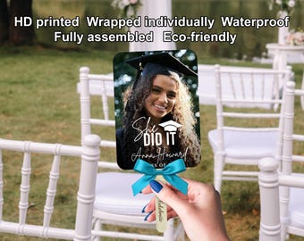 Può includere: Un ventaglio di laurea con la foto di una donna sorridente con il cappello di laurea. Il ventaglio presenta la scritta "She DID IT Anna Howard" ed è decorato con un fiocco turchese. Il ventaglio è tenuto davanti a uno sfondo sfocato all'aperto.