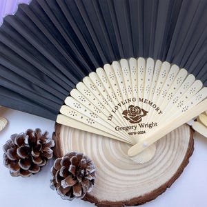 May include: Black and beige folding fan with the text "IN LOVING MEMORY" and the name "Gregory Wright 1979-2024". The fan is made of paper and wood. The fan is resting on a wooden surface with two pine cones.