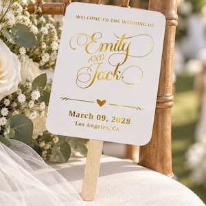 Könnte beinhalten: Ein weißer, rechteckiger Handfächer mit abgerundeten Ecken und einem Holzgriff. Der Fächer ist mit goldener Folienprägung versehen: WELCOME TO THE WEDDING OF Emily AND Jack, March 09, 2028, Los Angeles, CA, sowie einem kleinen goldenen Herzsymbol.
