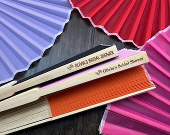 May include: A collection of colorful, folding fans in shades of purple, red, pink, and orange. The fans have wooden handles with the text "Olivia's Bridal Shower" engraved on them. The fans are open and fanned out.