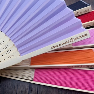 May include: A collection of colorful paper fans with wooden handles. The fans are in shades of purple, pink, orange, red, and black. Each fan is printed with the names "Olivia & Daniel" and the date "03.18.2026".
