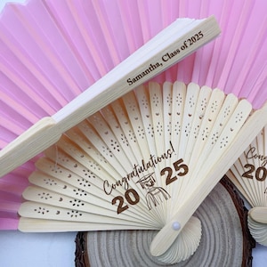 May include: Wooden graduation fans with the text "Congratulations! 2025" and "Samantha, Class of 2025". The fans are light beige with decorative cutouts and are partially fanned open. A pink paper fan is in the background.