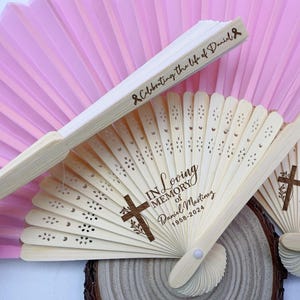 Puede incluir: Abanicos de mano de madera con diseños cortados con láser y texto. Un abanico dice "Celebrating the life of Daniel" con un símbolo de cinta. Otros abanicos tienen "In Loving Memory of Daniel Martinez 1958-2024" y una cruz.
