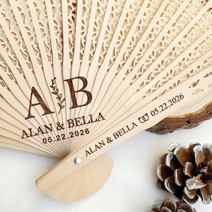 May include: A light-colored wooden fan with intricate cut-out designs. The fan is personalized with the names "ALAN & BELLA" and the date "05.22.2026". The fan is a wedding favor or decorative item.