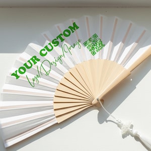 Può includere: Un ventaglio pieghevole con stecche in legno chiaro e pannelli in tessuto bianco. Il tessuto presenta il testo verde "YOUR CUSTOM Logo/Design/Image" e un codice QR verde. Una nappa bianca con piccole perline è fissata alla base del manico.