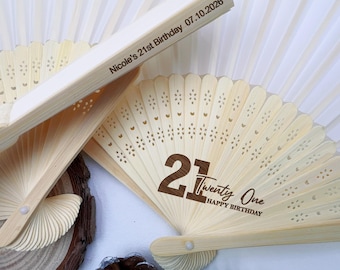 Op de afbeelding: Close-up van gepersonaliseerde houten waaiers. Eén waaier toont het nummer "21", de woorden "Twenty One" en "Happy Birthday". Een andere waaier heeft de tekst "Nicole's 21st Birthday 07.10.2026". De waaiers zijn lichtbeige met decoratieve uitsparingen.