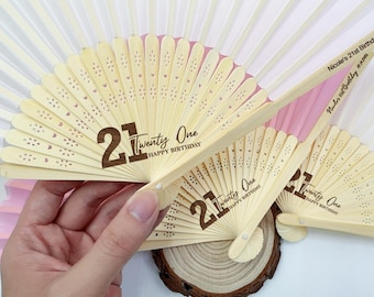 Op de afbeelding: Verschillende open, lichtgekleurde houten waaiers met een lasergesneden ontwerp. De waaiers hebben de tekst "21 Twenty One HAPPY BIRTHAY" in bruin gedrukt. Eén waaier wordt door een hand vastgehouden. De tekst "Nicole's 21st Birthday" staat op de handgreep.
