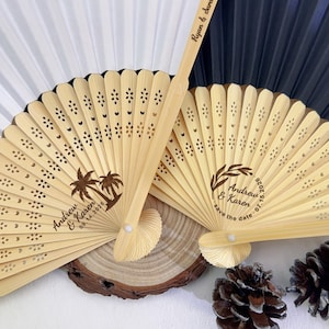 May include: Two wooden hand fans with intricate cut-out designs and personalized text. One fan features palm trees and the names "Andrew & Karen" with a date. The other fan has a wreath design and the names "Andrew & Karen" with a date. Wedding favors.