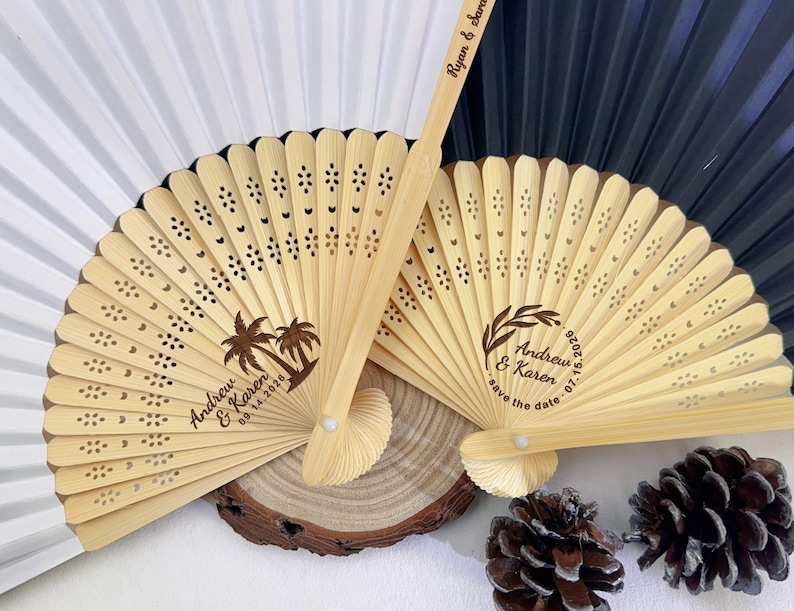May include: Two wooden hand fans with intricate cut-out designs and personalized text. One fan features palm trees and the names "Andrew & Karen" with a date. The other fan has a wreath design and the names "Andrew & Karen" with a date. Wedding favors.