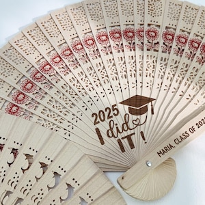 Könnte beinhalten: Holz-Abschlussfächer mit lasergeschnittenen Blumen- und geometrischen Mustern. Der Fächer trägt den Text "2025 I did it!" und "Maria, Klasse von 2025". Der Fächer ist hellbeige mit roten floralen Akzenten.