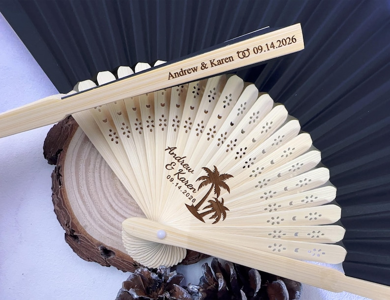 May include: A light-colored wooden fan with laser-cut floral designs and the names "Andrew & Karen" along with the date "09.14.2026". The fan features a palm tree graphic and is partially open, with a black fan in the background.