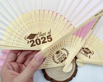 May include: Several open, light-colored wooden fans with laser-cut designs. The fans have the text "Class of 2025" and "Finally 2025" printed on them. One fan has the names "Maria, Class of 2025" and "Samantha, Class of 2025" printed on it. The fans are on a wooden surface.