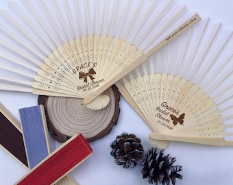 May include: A collection of decorative fans, some open and some closed, in various colors. Two fans are open, displaying the text "Grace's Bridal Shower 01.23.2026". The fans are made of wood and paper. Additional fans are visible in red, blue, and brown.