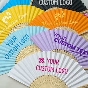 May include: A collection of colorful, personalized paper fans. The fans are in various colors, including yellow, blue, orange, purple, and white. Each fan has the text "Your Custom Logo" or "Your Custom Text" printed on it. The fans have a wooden handle.