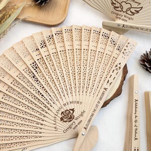 Op de afbeelding: Lichtbruine houten waaiers met ingewikkelde uitgesneden patronen en de tekst "In Loving Memory Gregory Wright 1979-2024 Celebrating the Life of Gregory". De waaiers hebben een bloemenpatroon. Sommige zijn open, andere gesloten.