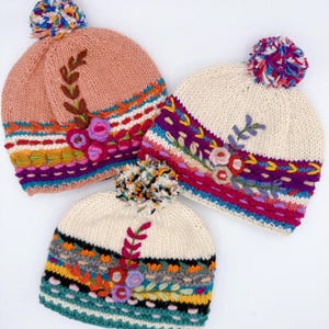 Peut inclure: Trois bonnets en tricot avec pompons. L'un est pêche, deux sont crème. Chacun présente des bandes horizontales brodées colorées, incluant des motifs floraux avec des feuilles et des tiges dans des tons de rose, violet, orange, jaune et bleu sarcelle.