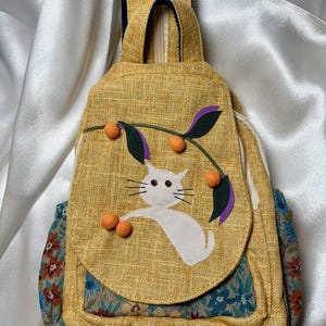 Puede incluir: Una mochila de lona amarilla con un diseño de gato blanco y fruta naranja. La mochila tiene un patrón floral azul y verde en la parte inferior.