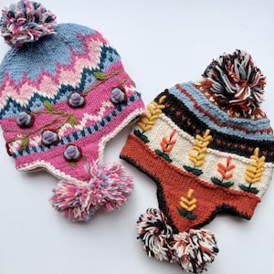May include: Two colorful knit hats with ear flaps and pom-poms. One hat is pink and blue with floral embroidery and a zig-zag pattern. The other hat is cream, orange, and black with a wheat-like design.