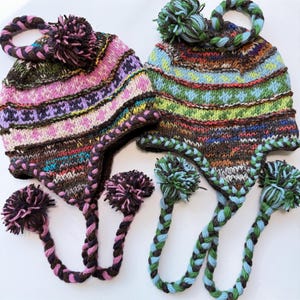 May include: Two knit earflap hats with braided tassels and pom-poms. The left hat features pink, purple, and brown geometric patterns. The right hat displays blue, green, and brown geometric patterns. Both hats have braided ties.
