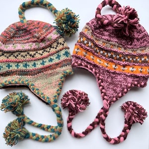 Könnte beinhalten: Zwei gestrickte Mützen mit Ohrenklappen, geflochtenen Bändern und Bommeln. Die linke Mütze hat ein mehrfarbiges Muster aus rosa, blaugrünem und beigem Garn. Die rechte Mütze zeigt ein lebhaftes Design in Magenta-, Orange- und Brauntönen.