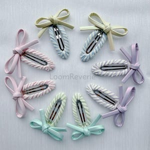 May include: Eight hair clips arranged in a circle, each with a fabric-covered clip and a bow. The clips are in pastel shades of pink, blue, green, and lilac. The text "LoomReverie" is visible. These are ideal for adding a touch of colour.
