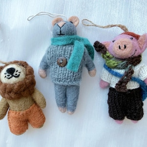 Puede incluir: Tres adornos de animales de fieltro hechos a mano: un león con pantalones cortos naranjas, un ratón gris con una bufanda turquesa y un cerdo rosa con bufanda y suéter. Cada adorno tiene una cuerda para colgar.
