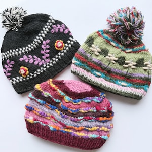 May include: Three colorful knit beanies with pom-poms. One is black with white and pink floral embroidery and a white pom-pom. Another is green with white and tan accents and a multi-colored pom-pom. The third is burgundy with colorful wavy stripes.