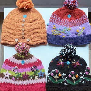 May include: Four knitted beanies with pom-poms. Top left is orange with wooden buttons. Top right is red, purple, and blue with floral embroidery. Bottom left is pink, green, and brown with floral patterns. Bottom right is black with floral embroidery.