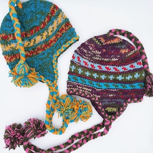 May include: Two colorful knitted hats with ear flaps and pom-poms. One hat is blue with orange, yellow, and red accents. The other is purple with blue, green, and red details. Braided ties and pom-poms complete the design.