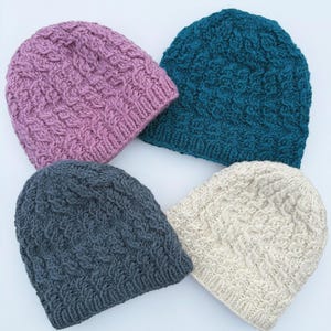 May include: Four knitted beanies in shades of purple, teal, gray, and cream. Each hat features a textured cable knit pattern. The beanies are arranged in a square formation, showcasing their different colors and textures.