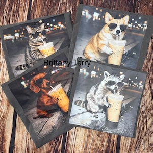 Puede incluir: Cuatro posavasos cuadrados de color gris oscuro con ilustraciones de animales. Se representan un gato, un corgi, un perro salchicha y un mapache con gafas de sol y bebiendo caf&eacute; helado. El texto "Brittany Terry" es visible.