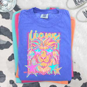 May include: A purple short-sleeve t-shirt featuring a graphic of a lion wearing star-shaped sunglasses. The text "Lions" appears above the lion, and "Brittany" is printed below. The design uses bright neon colors including pink, yellow, and blue.