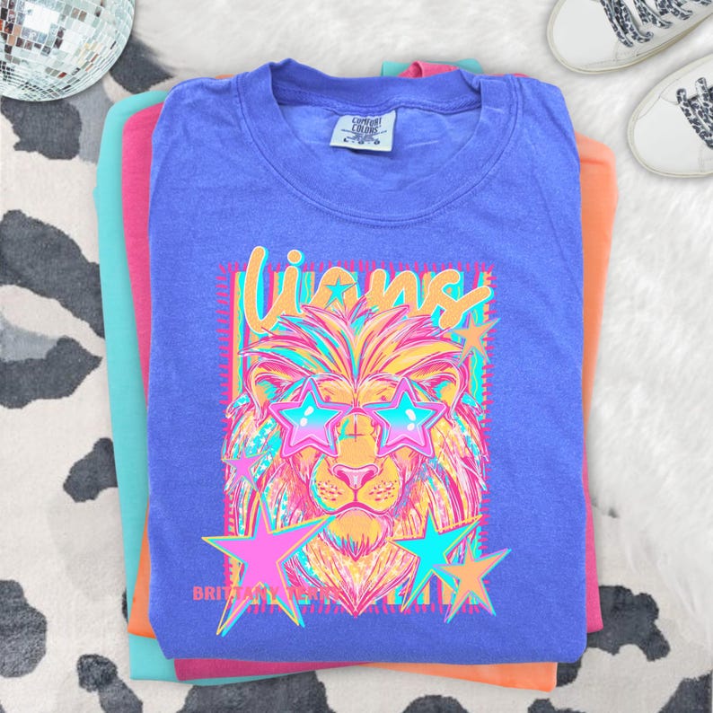 May include: A purple short-sleeve t-shirt featuring a graphic of a lion wearing star-shaped sunglasses. The text "Lions" appears above the lion, and "Brittany" is printed below. The design uses bright neon colors including pink, yellow, and blue.