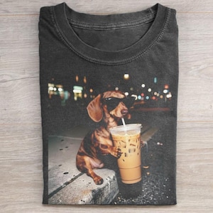 Puede incluir: Camiseta gris oscuro con un perro salchicha con gafas de sol bebiendo café helado. El perro está sentado en una repisa de hormigón con un fondo urbano borroso. La camiseta es de material suave.