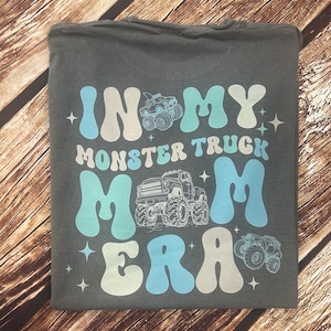 Pu&ograve; includere: T-shirt grigio scuro con la scritta "IN MY MONSTER TRUCK MOM ERA" in bianco e azzurro. Il design include illustrazioni di monster truck e stelle. La maglietta &egrave; piegata su una superficie di legno.